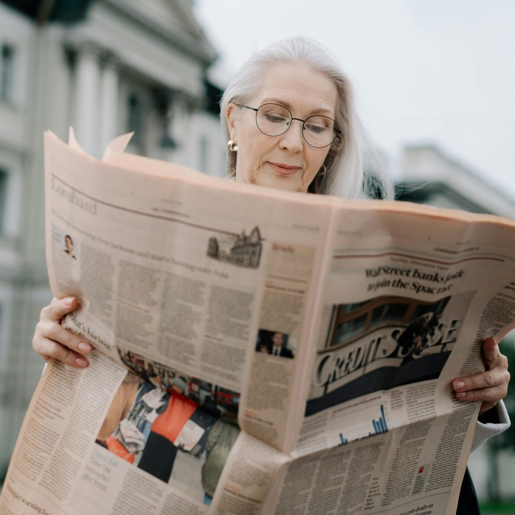 Woman with grey hair reading newspaper outside of building.